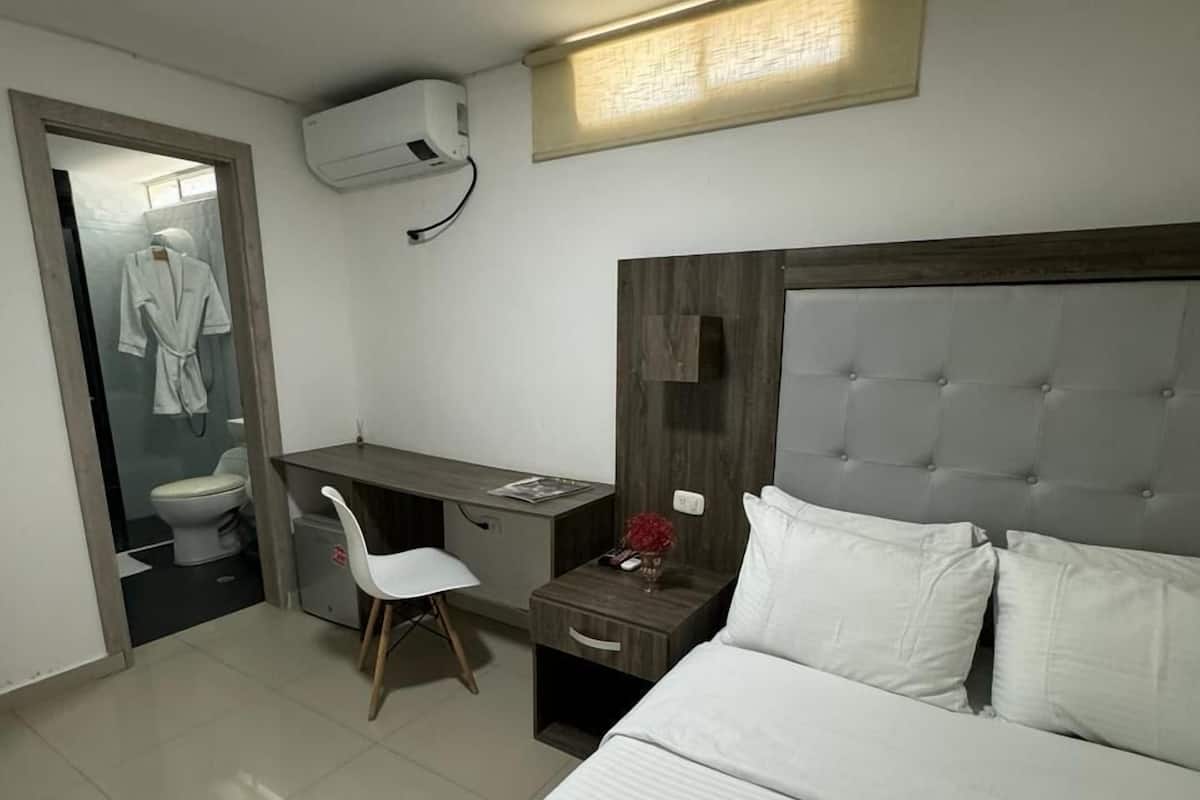 Une chambre d’hôtel avec un lit, un bureau, une chaise et une salle de bain visible à travers une porte ouverte.