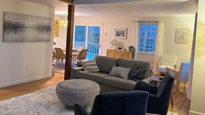A spacious living room with a high ceiling, a wooden staircase, a large sofa, and a dining area in the background.
