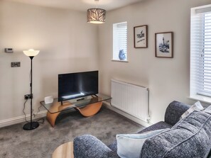 A living room with a flat-screen TV on a glass stand, a floor lamp, a radiator, and a sofa with patterned cushions.