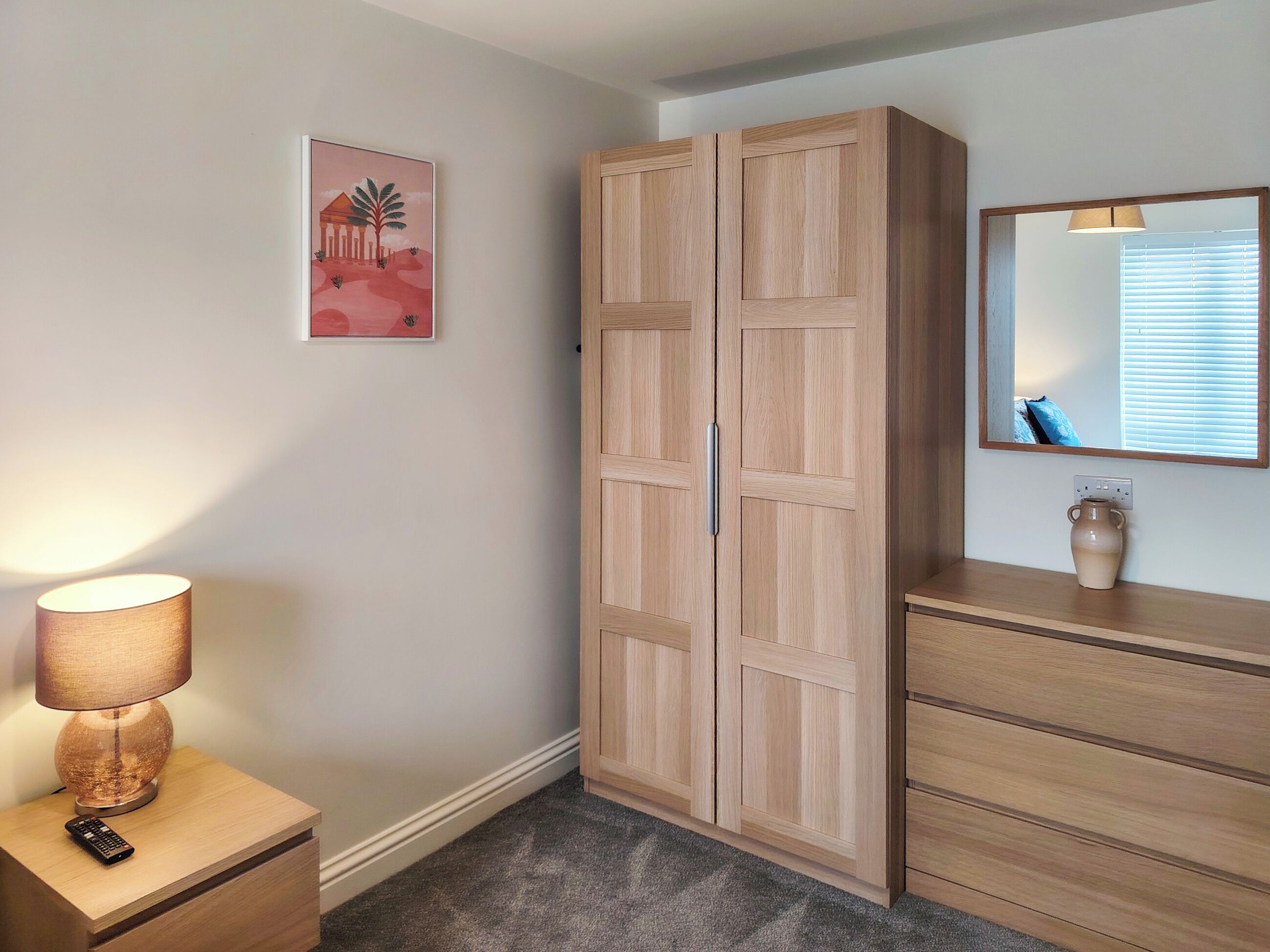 A room with a wardrobe, a bedside table with a lamp, a vase, and a painting on the wall.