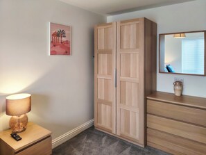 A room with a wardrobe, a bedside table with a lamp, a vase, and a painting on the wall.