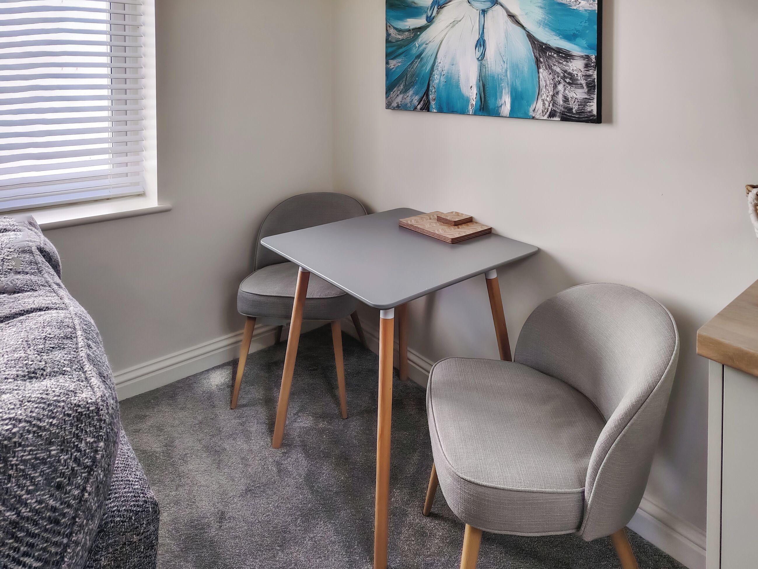 A room with a grey carpet, a grey table with a herringbone-patterned book, two grey chairs, a painting on the wall, and a window with blinds.