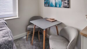 A room with a grey carpet, a grey table with a herringbone-patterned book, two grey chairs, a painting on the wall, and a window with blinds.