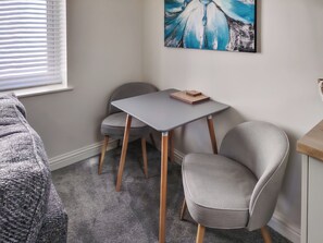 A room with a grey carpet, a grey table with a herringbone-patterned book, two grey chairs, a painting on the wall, and a window with blinds.