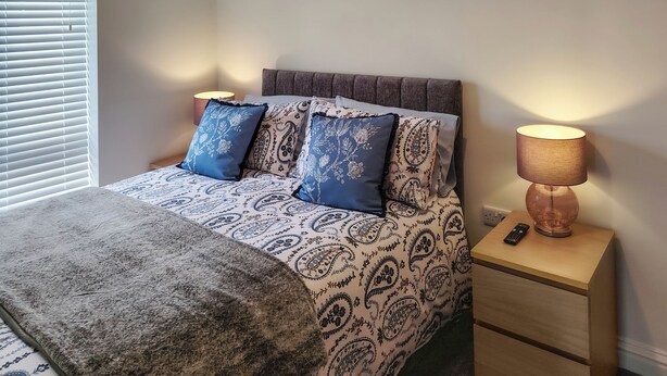 A bedroom with a bed, patterned bedding, a bedside lamp, a remote control, and a window with blinds.