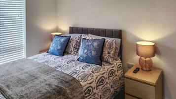 A bedroom with a bed, patterned bedding, a bedside lamp, a remote control, and a window with blinds.
