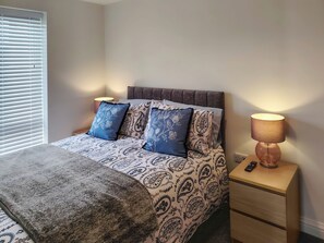 A bedroom with a bed, patterned bedding, a bedside lamp, a remote control, and a window with blinds. - 1 bedroom accommodation in Filey (Filey)