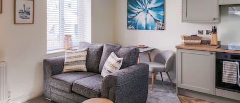 Uma sala de estar aconchegante com um sofá cinza, uma pequena mesa redonda, uma poltrona cinza, uma área de cozinha com armários e um grande quadro floral azul na parede.