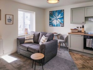 A cozy living room with a grey sofa, a small round table, a grey chair, a kitchen area with cabinets, and a large blue floral painting on the wall. - 1 bedroom accommodation in Filey (Filey)
