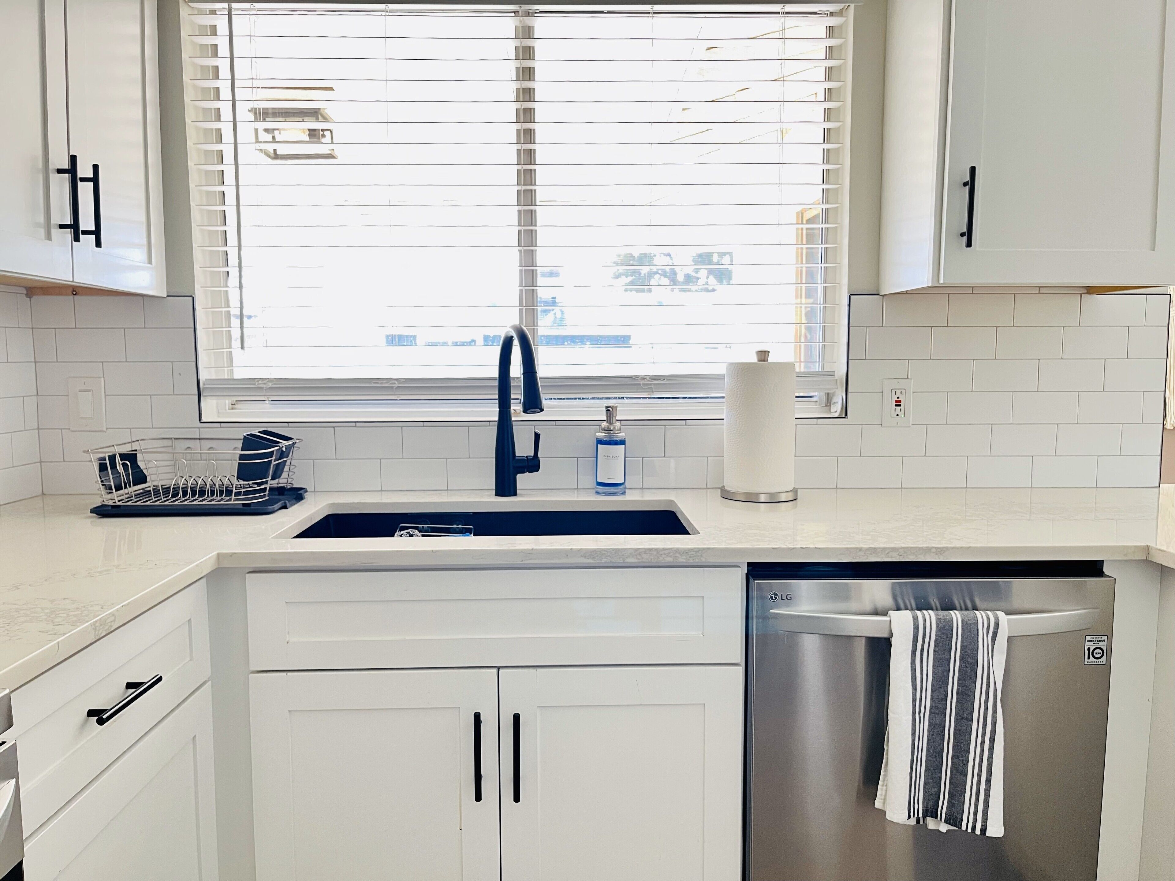 A modern kitchen with white cabinets, a stainless steel dishwasher, and a window with blinds.
