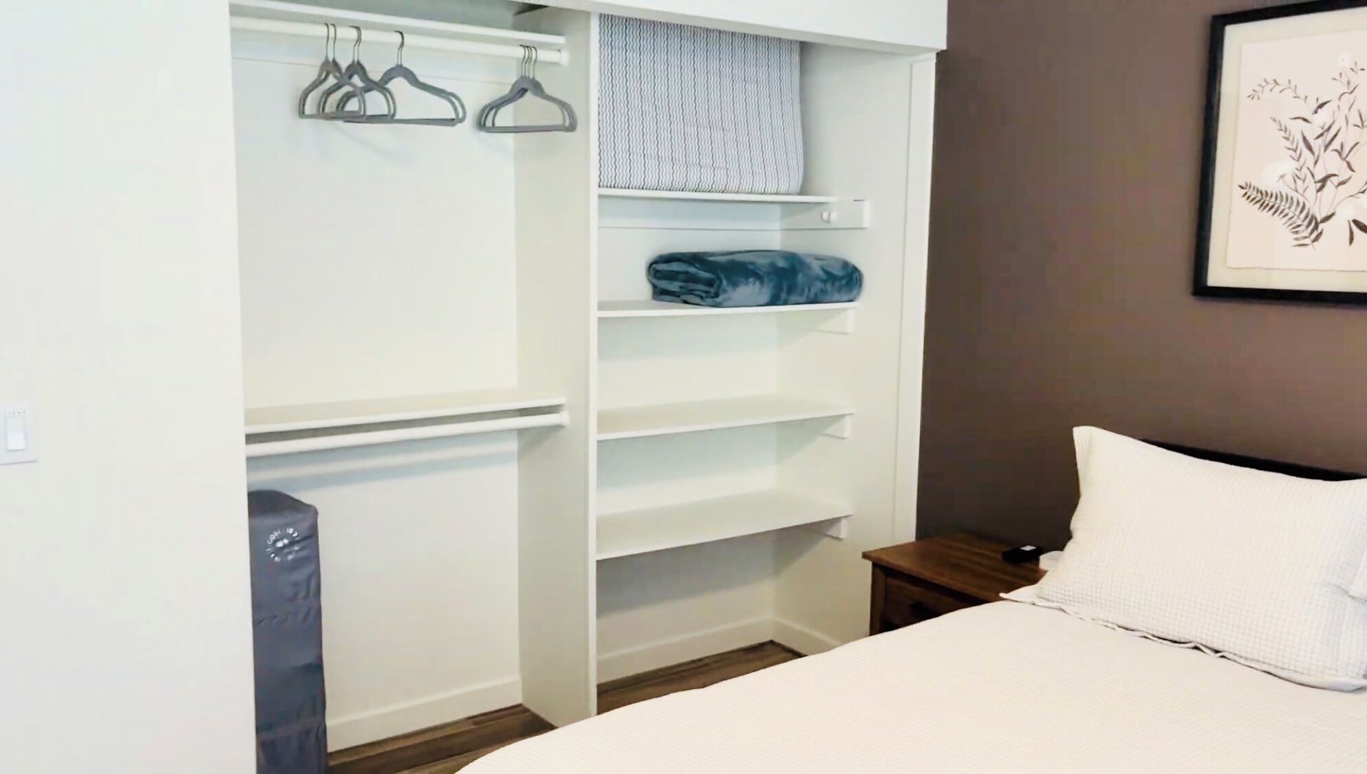 A bedroom with a bed, a nightstand, a wardrobe with hangers, and a framed picture on the wall.