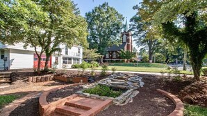 A landscaped garden with a brick pathway leading to a circular stone feature, surrounded by trees and a building with a steeple in the background. - Charming 4-bedroom house with AC and WiFi in delightful Troutman (Troutman)