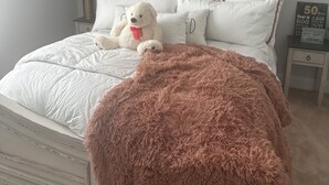 Ein ordentlich gemachtes Bett mit einer flauschigen, rosa Decke, weißer Bettwäsche und einem Teddybären. Ein Nachttisch mit Lampe und einer kleinen Schrifttafel steht daneben.