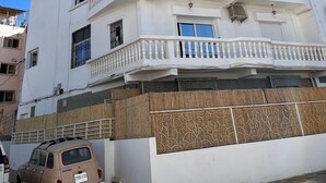 A multi-story white building with balconies, a wooden fence, and a parked car. - Paddle Out Morocco (Tamraght)