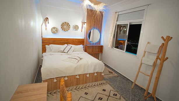 A neatly arranged bedroom with a wooden headboard, a bed with white bedding, a small rug, and a wooden ladder against the wall. - Paddle Out Morocco (Tamraght)