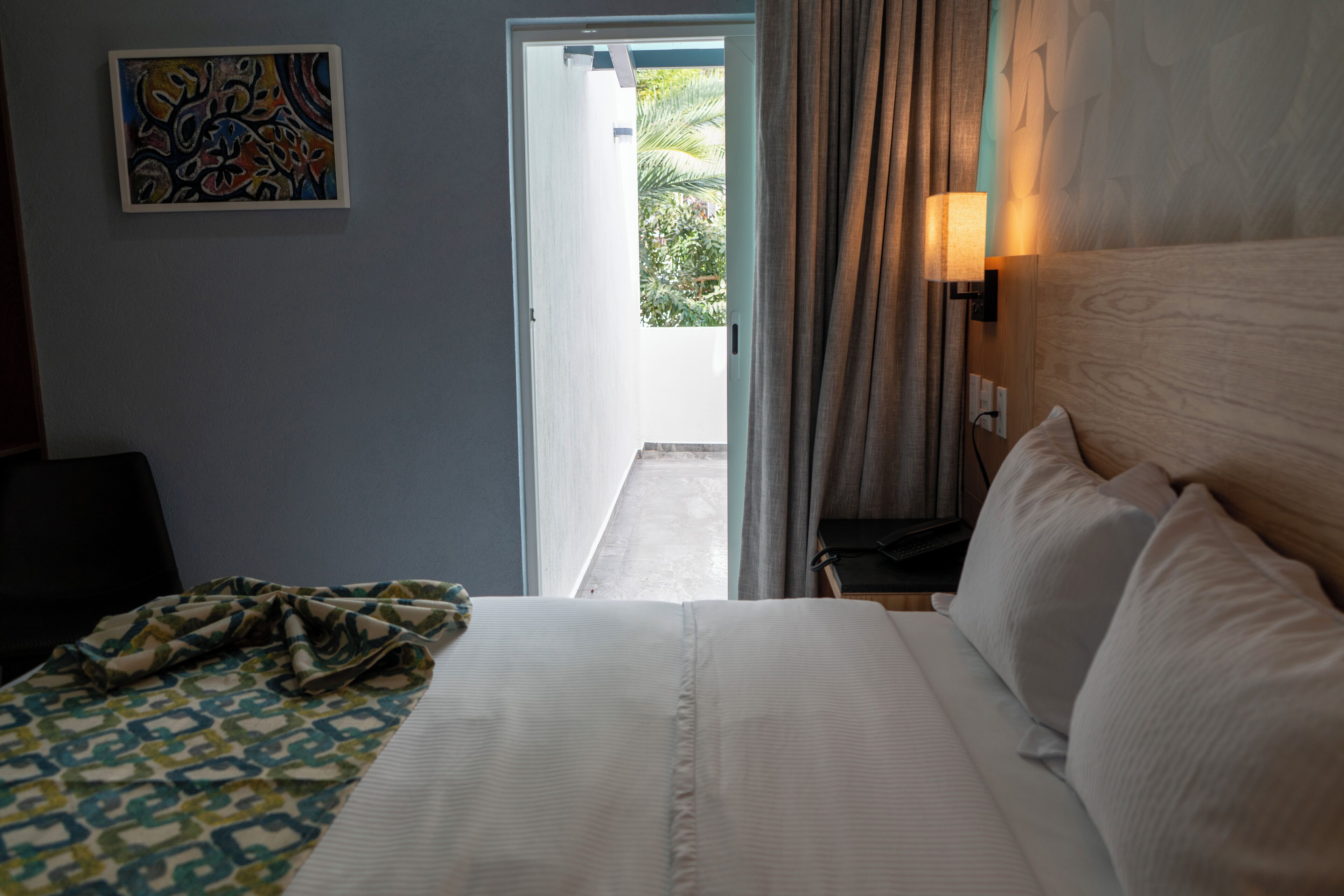 A hotel room with a bed, a nightstand, a lamp, a painting on the wall, and an open door leading to an outdoor area.