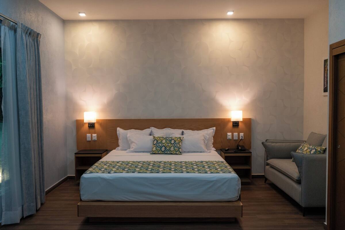 A hotel room with a bed, two bedside tables, a nightstand, a sofa, and a window with curtains.