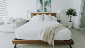 Une chambre moderne avec un lit en bois, du linge de lit blanc, un fauteuil blanc et des œuvres d’art abstraites accrochées au mur.