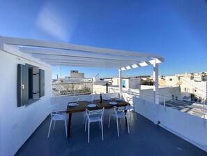 En tagterrasse med et træbord dækket til spisning, omgivet af hvide stole, under en hvid pergola.