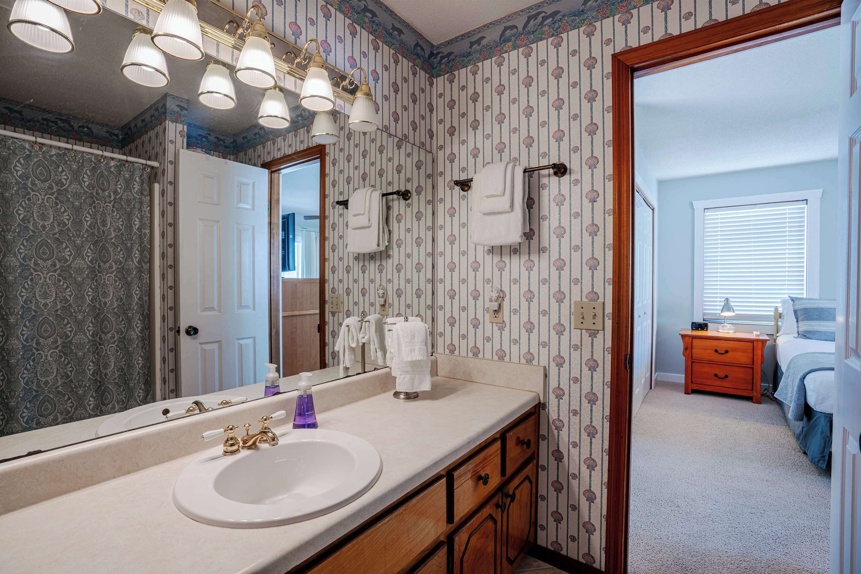 A bathroom with a large mirror, a sink, and a vanity with drawers. There is a towel rack above the sink and a window with blinds in the background.