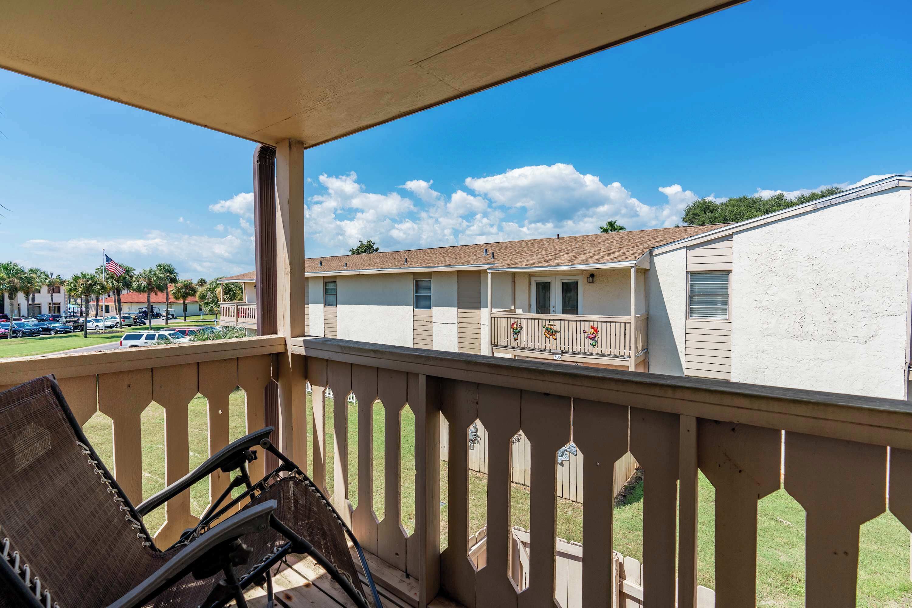 A motel with a covered balcony, outdoor seating, and a view of the parking lot and neighboring buildings.