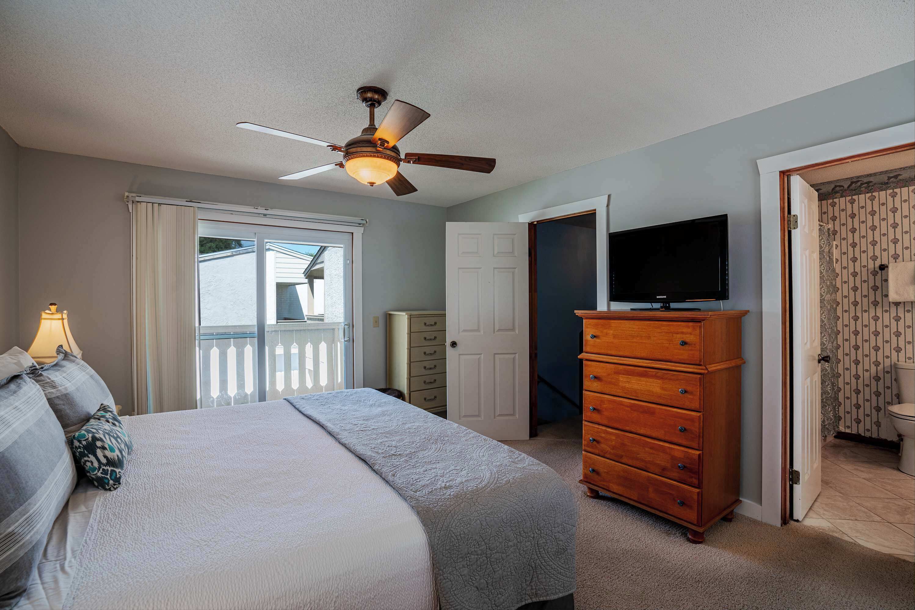 A bedroom with a bed, a dresser, a ceiling fan, a television, and a bathroom with a toilet and shower.