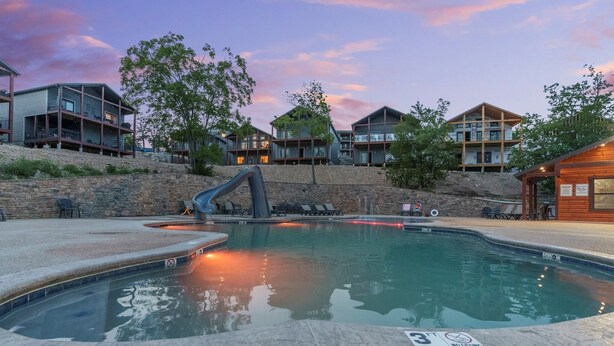 A swimming pool with a hot tub, surrounded by modern buildings and a stone wall. - Poolside | Lake Views | Game Room | 2-Min to SDC (Branson)