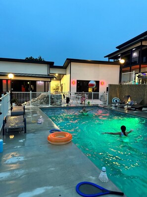 Zona de piscina con sillas de descanso y una mesa junto a la piscina, rodeada de edificios de diseño moderno.