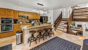 Cocina moderna con armarios de madera, encimera de granito y refrigerador de acero inoxidable. Cuenta con una zona de bar con taburetes y una escalera que sube a una sala de estar en un entrepiso.