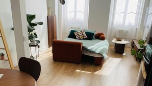 Et moderne, veloplyst rum med trægulv, en seng med grønt sengetøj og orange puder, et lille rundt bord med en skål og en potteplante.