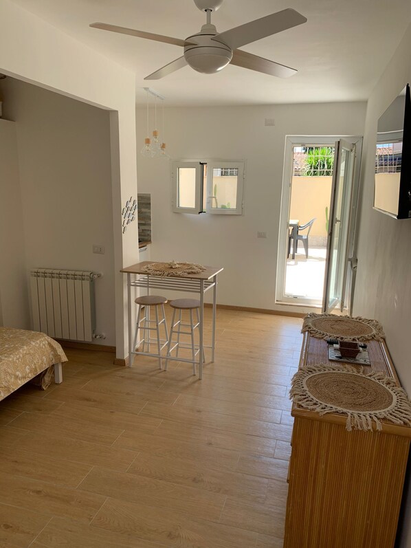 A bedroom with a bed, a table with two stools, a ceiling fan, a radiator, and a TV mounted on the wall.