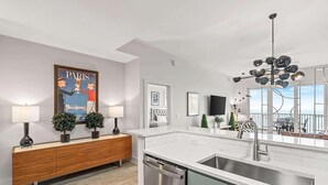 A modern kitchen with a white countertop, stainless steel sink, and a dishwasher. There is a wooden console table with two lamps and small potted plants. A large poster of Paris is on the wall, and a TV is mounted above the console table. - Salt and Shore Escape Sleeps 7 (Panama City Beach)