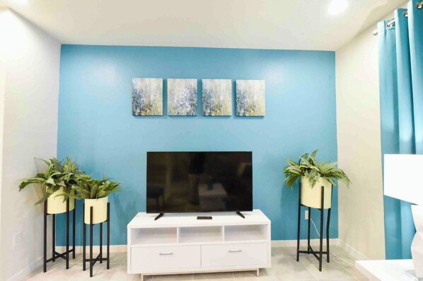 Una sala de estar con un televisor de pantalla plana grande sobre un mueble blanco, flanqueado por dos plantas en macetas sobre pedestales negros.