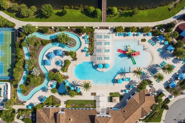 An aerial view of a resort with a central swimming pool, multiple lounge areas, and a sports court.