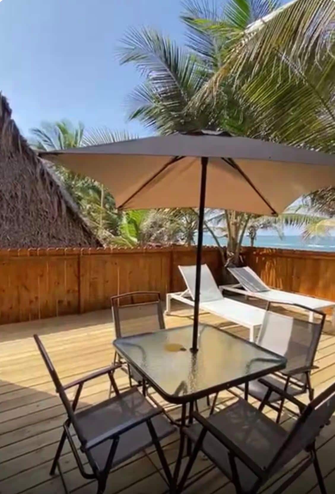 Eine Holzterrasse mit einem Glastisch und Stühlen, einem großen Sonnenschirm und Liegestühlen.