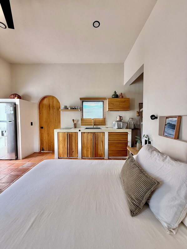 A bedroom with a large bed, a wooden cabinet, a small kitchen area with a refrigerator, and a window with blinds.