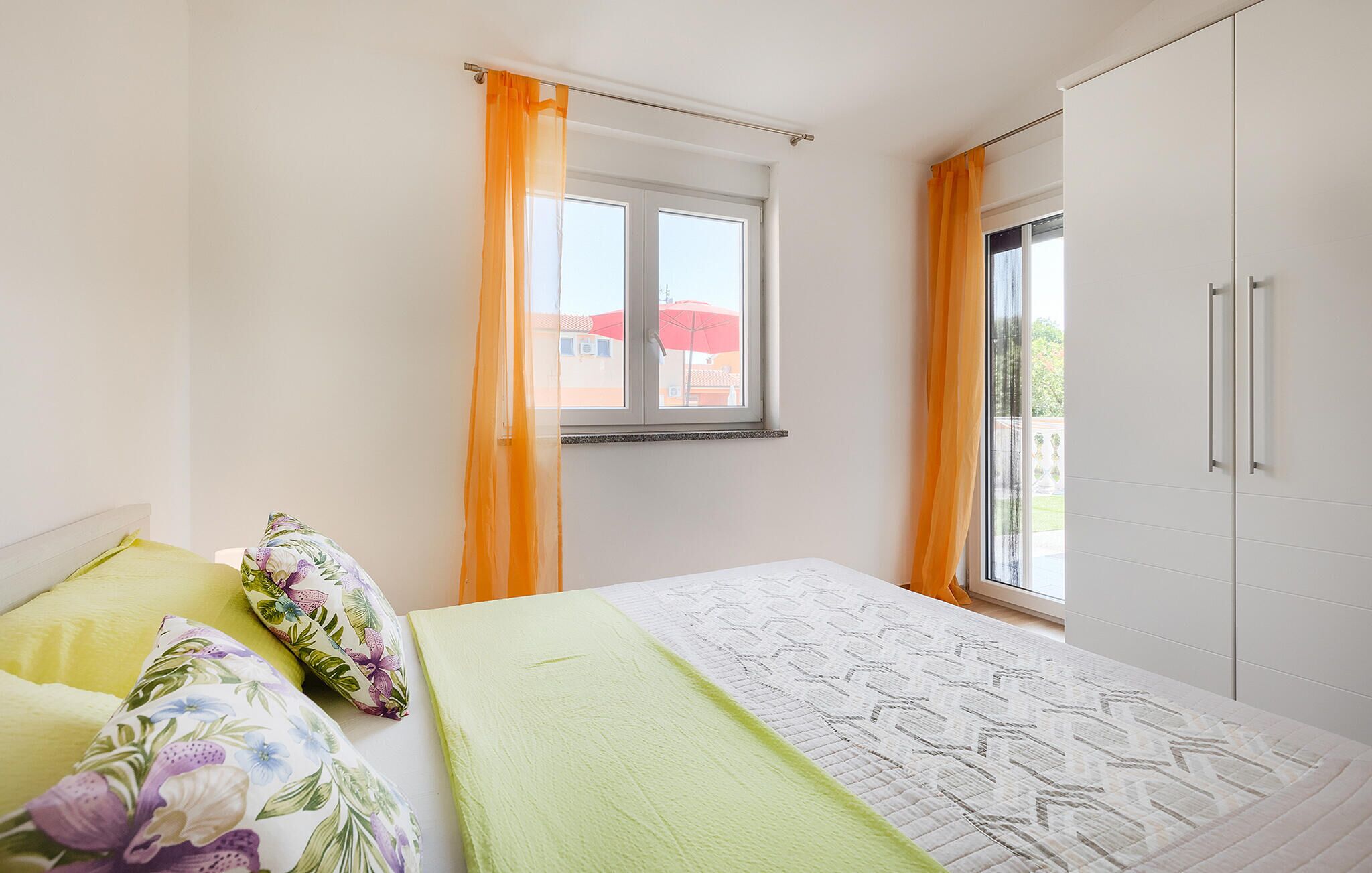 Une chambre à coucher avec un lit, une fenêtre ornée de rideaux orange, une vue sur un bâtiment rouge à l’extérieur et une armoire blanche.