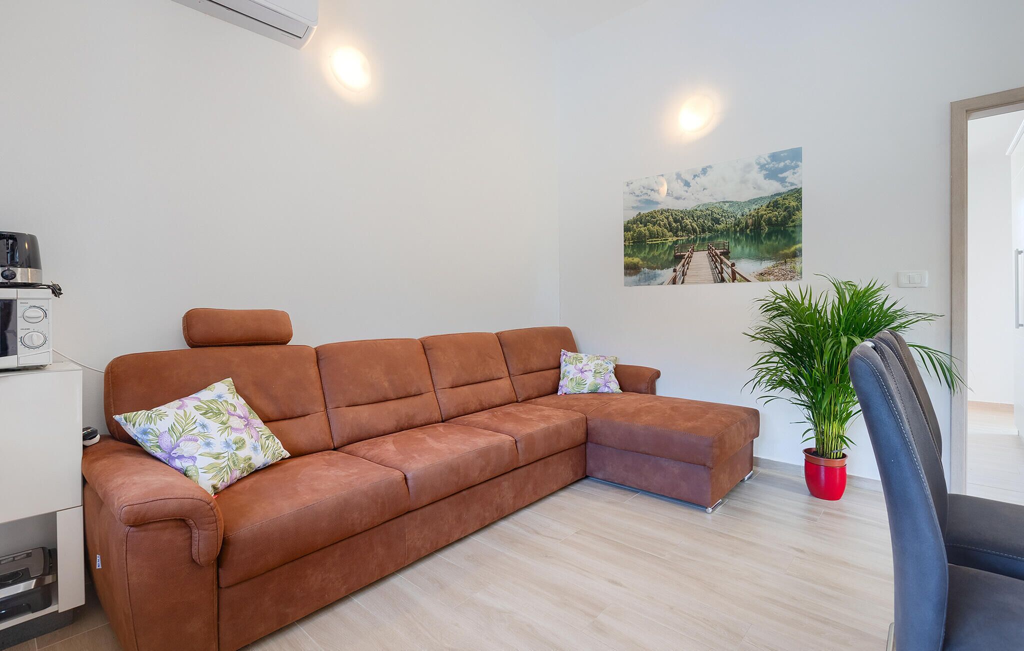 A modern living room with a brown leather sofa, a small table with a kettle, a potted plant, and a painting of a lake.