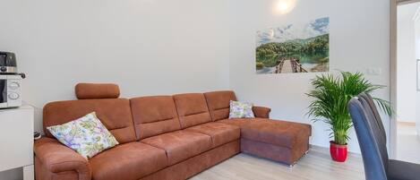 A modern living room with a brown leather sofa, a small table with a kettle, a potted plant, and a painting of a lake.