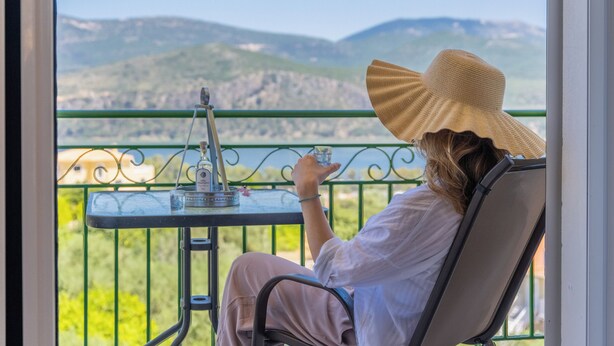 Una persona sentada en una silla de balcón, sosteniendo un vaso de agua, con vistas a montañas y vegetación al fondo.