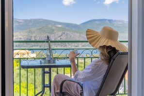 A person seated on a balcony chair, holding a glass of water, with a view of mountains and greenery in the background.