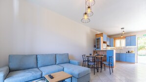 Una sala moderna con un sofá seccional azul claro, una pequeña mesa de madera y un control remoto sobre la mesa. Hay una zona de comedor con una mesa y sillas, y una cocina con armarios azules y un ventanal.