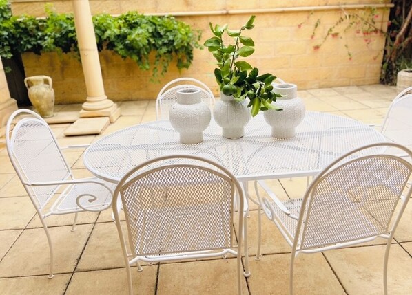 Mesa redonda branca de metal com quatro cadeiras iguais, rodeada por três vasos brancos de cerâmica com plantas verdes.