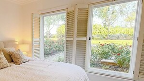 Une chambre à coucher avec un grand lit, une table de chevet, une lampe et une vue sur un jardin, les volets étant ouverts.
