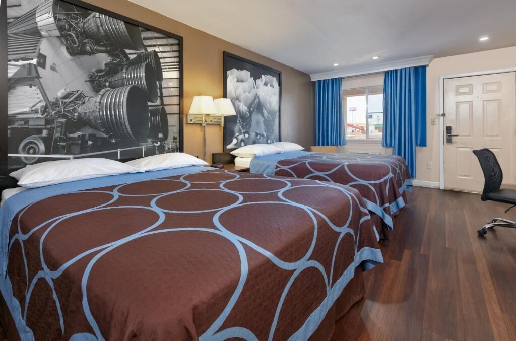 A hotel room with two beds, a large poster of a rocket launch, a desk with a lamp, and a view of the outside through a door.