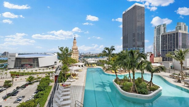 A resort pool area with a view of a tall building, palm trees, and a modern building with a sign. - PH 51 - 1BD at NEW Worldcenter area + free parking (Miami)