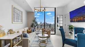 A modern living room with a sofa, coffee table, and chairs, a dining area with a table and shelves, and a large window with a city view. - PH 51 - 1BD at NEW Worldcenter area + free parking (Miami)