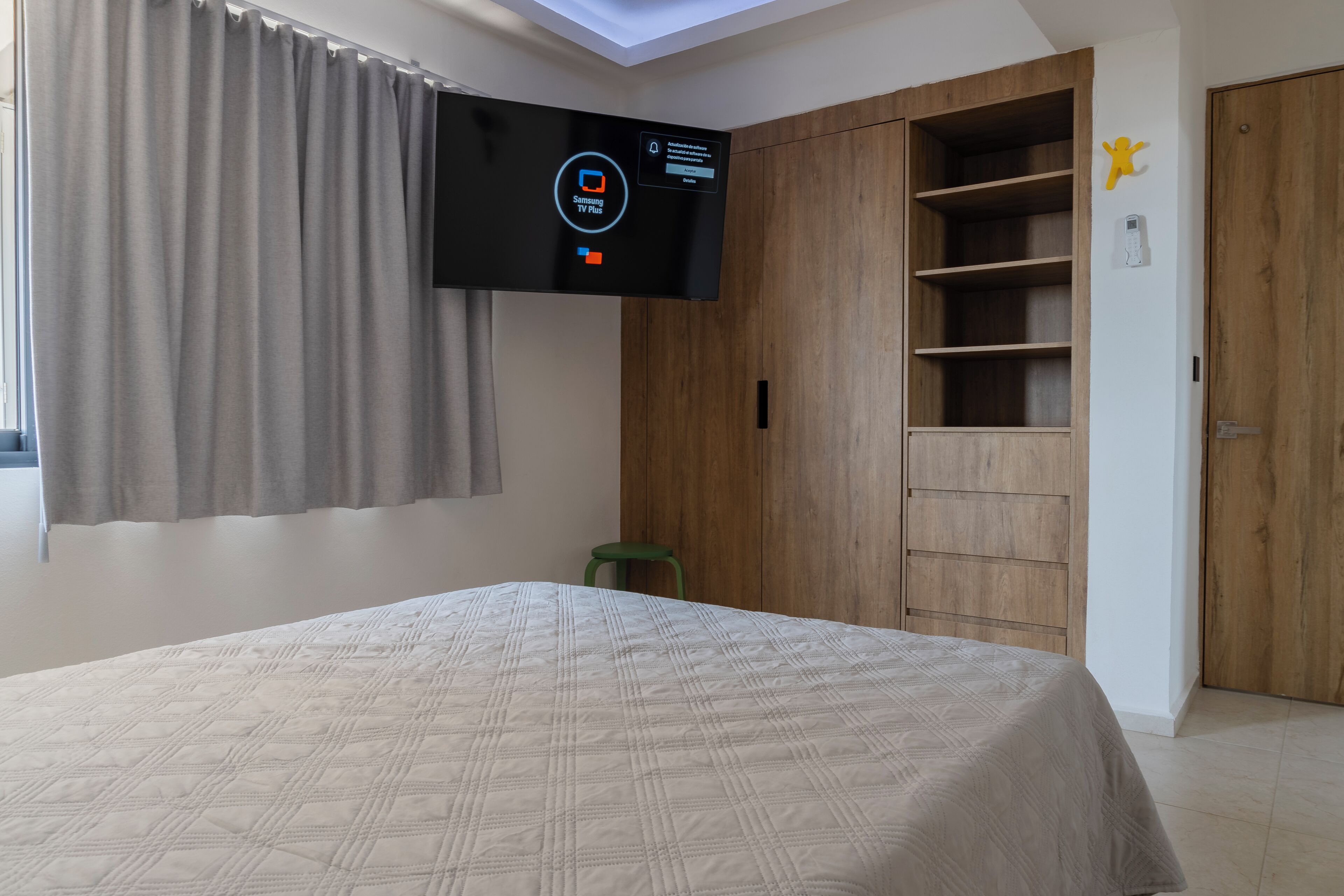 A bedroom with a bed, a TV mounted on the wall, a wooden wardrobe, and a green stool.