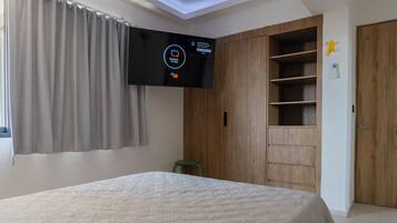 A bedroom with a bed, a TV mounted on the wall, a wooden wardrobe, and a green stool.