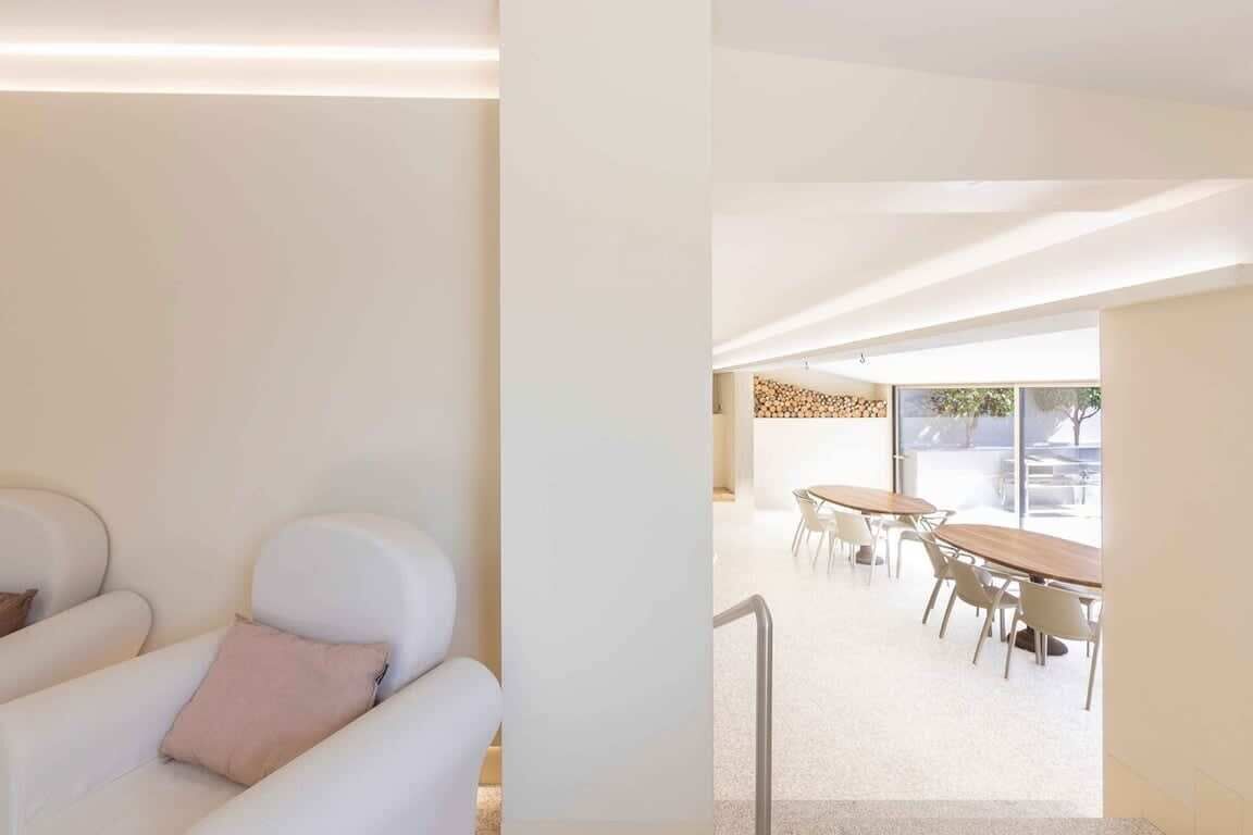 Un interior modern amb un sofà blanc, una zona de menjador amb taula i cadires de fusta, i una vista a través d'una porta de vidre.